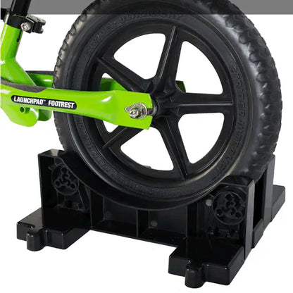 Bright green launchpad footrest with black tire and wheel on sturdy black plastic stand for strider bike storage base