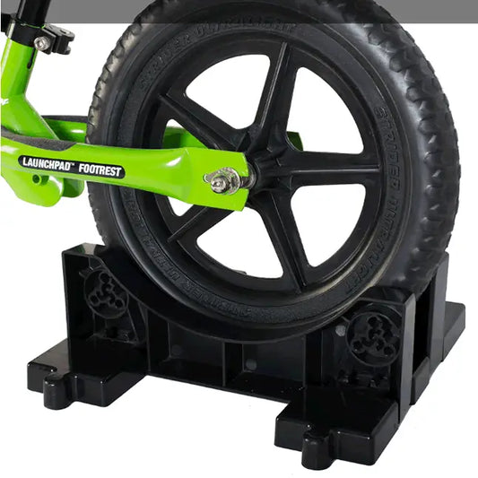 Bright green launchpad footrest with black tire and wheel on sturdy black plastic stand for strider bike storage base
